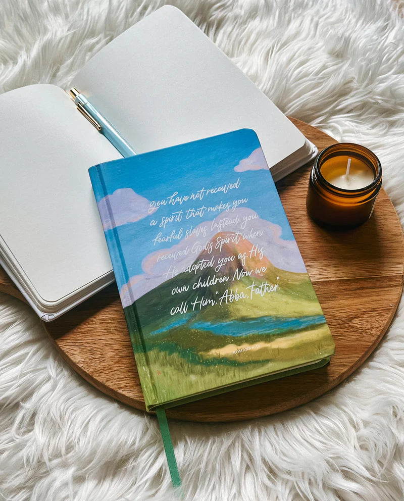 Hardcover journal with Romans 8 verse and painted sky design by Project J placed on wooden tray beside open notebook and candle.