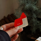 A hand holding a colorful acrylic magnet shaped like a house with the word "Home" displayed prominently, set against a softly blurred background of greenery.