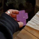A hand holding a vibrant purple acrylic magnet shaped like a cross with the name "Jesus" inscribed on it, set against a softly blurred background of greenery.