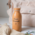 Mustard-colored milk bottle tumbler with the handwritten phrase "always hallelujah" and a smiley face, set on a light surface with a beige bag and cozy sweater in the background.