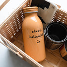 Mustard-colored milk bottle tumbler with the handwritten phrase "always hallelujah" and a smiley face, shown in a wooden gift basket.