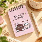 Always Loved pink spiral notepad on a sunlit desk beside coffee and pen