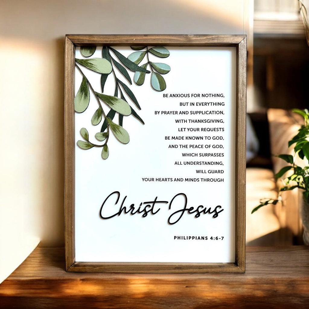 Framed wall art with the Bible verse Philippians 4:6-7, featuring greenery and the phrase 'Christ Jesus' in script font, displayed in a warmly lit room.