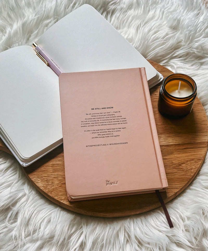 Blush pink hardcover journal with printed message on back cover by Project J placed on wooden tray beside open notebook and candle.