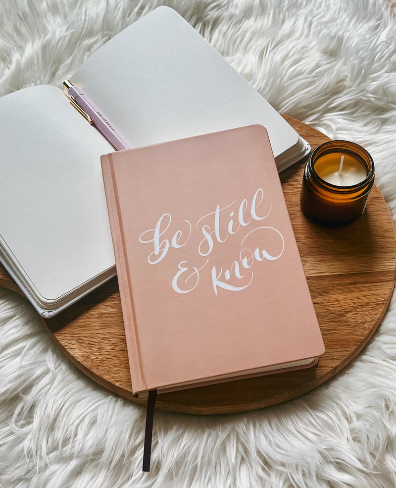 Blush pink hardcover journal with “Be still & know” text by Project J placed on wooden tray beside open notebook and candle.
