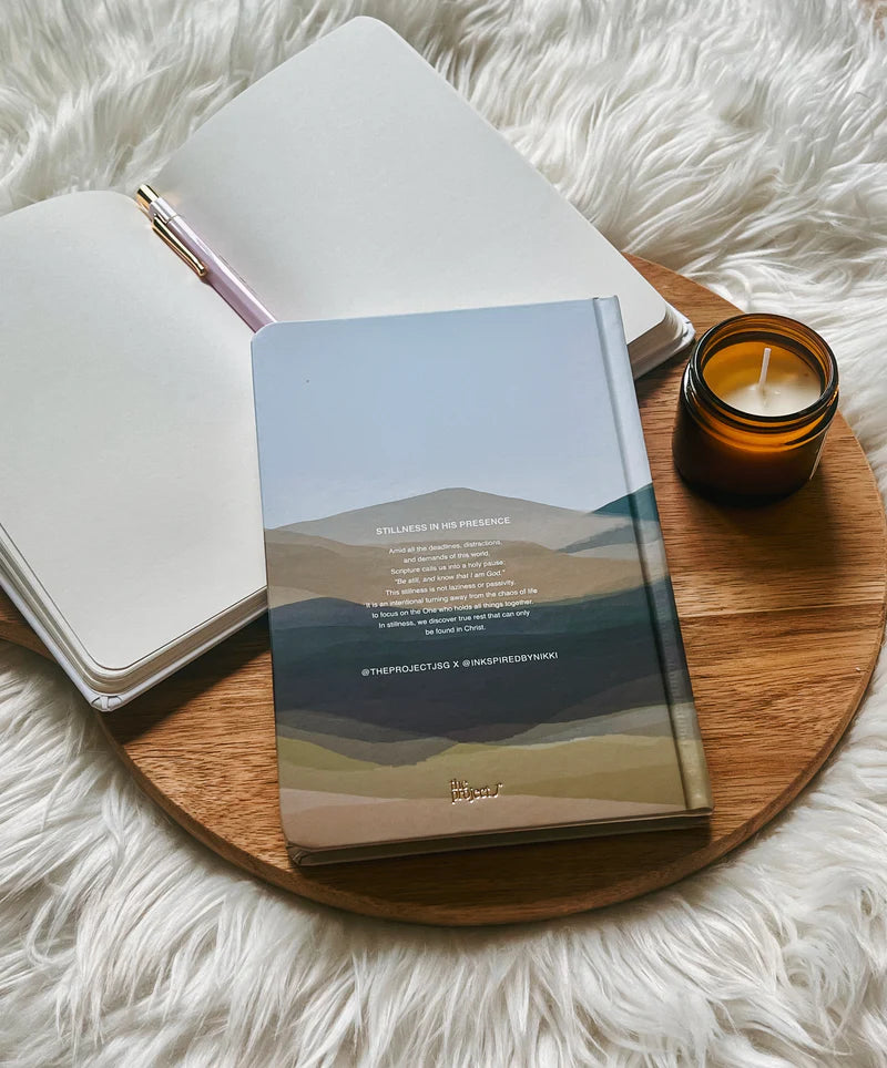 Hardcover journal with printed reflection text on back cover by Project J placed on wooden tray beside open notebook and candle.