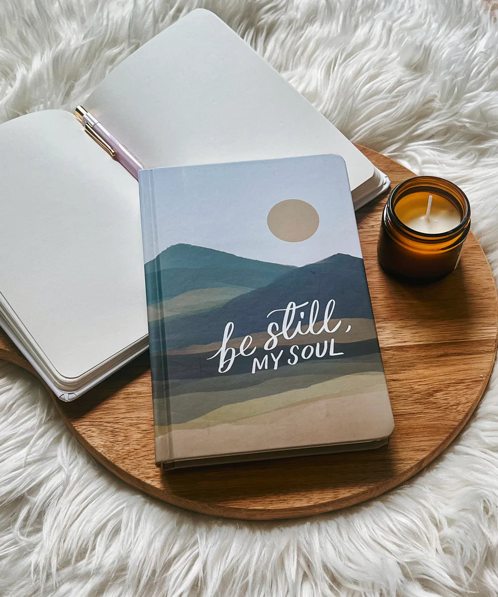Hardcover journal with “Be still, my soul” lettering design by Project J placed on wooden tray beside open notebook and candle.
