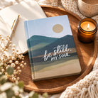 Be Still, My Soul Christian journal styled on a wooden tray with an open notebook, candle and soft neutral textiles.