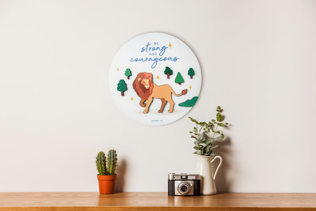 3D wooden Christian wall decor featuring the phrase 'Be Strong and Courageous,' with a lion and trees, inspired by the Bible verse Joshua 1:9, displayed above a shelf with plants and a vintage camera.