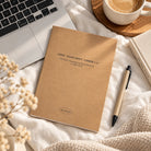 Because He First Loved Us 1 John 4:19 A5 kraft notebook lying on cream bedding beside a laptop and coffee