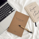 Flat lay of “Because He First Loved Us” Christian notebook featuring 1 John 4:19, with a pen and laptop, ideal for faith journaling and encouragement gifts from The Commandment Co Singapore.