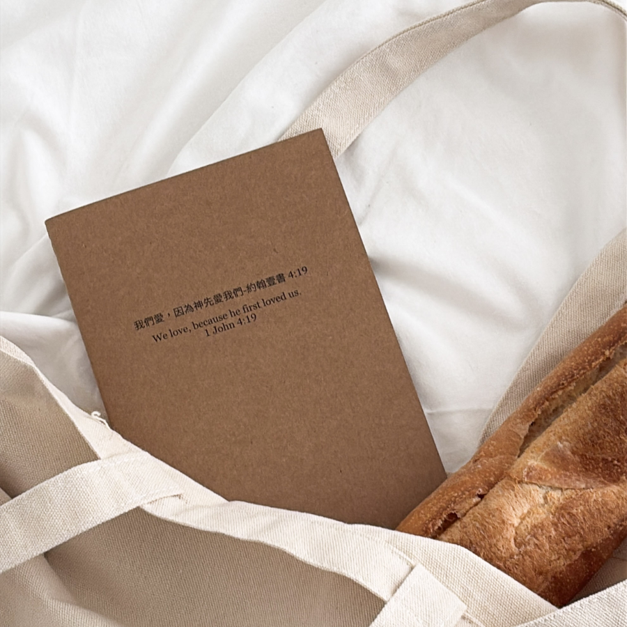 Because He First Loved Us notebook with Bible verse 1 John 4:19, pen, and laptop on a white desk, perfect Christian gift from The Commandment Co Singapore.