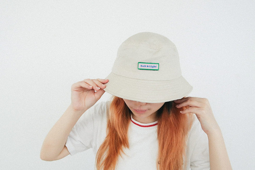 A person wearing a beige bucket hat with a green and white velcro patch reading "Salt & Light," highlighting a customizable faith-inspired streetwear accessory.