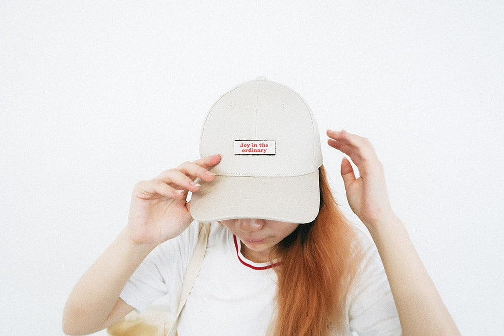 A person wearing a beige cap with a red and white velcro patch that reads "Joy in the ordinary," showcasing faith-based and customizable casual streetwear.