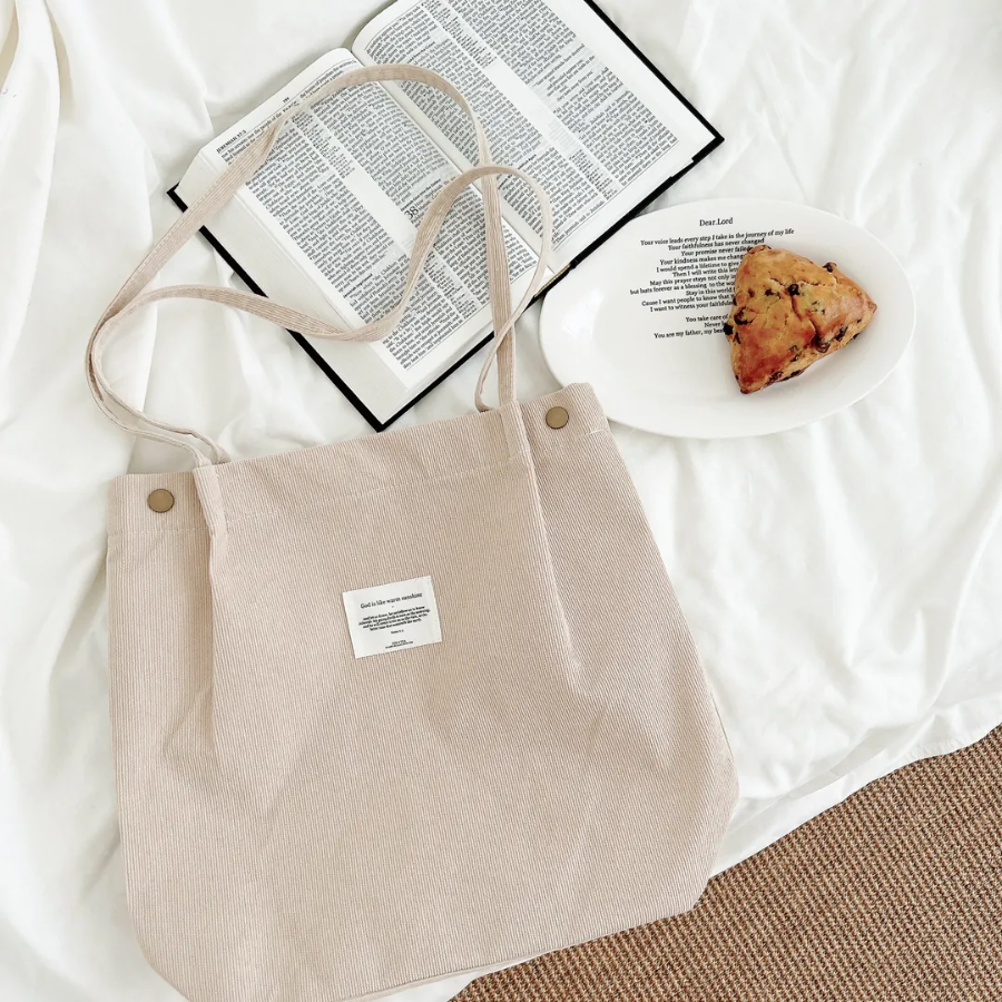 Beige corduroy shoulder bag with an inspirational patch, placed on a cozy white bed next to an open Bible and a plate with a scone, reflecting simple happiness and faith-inspired living.