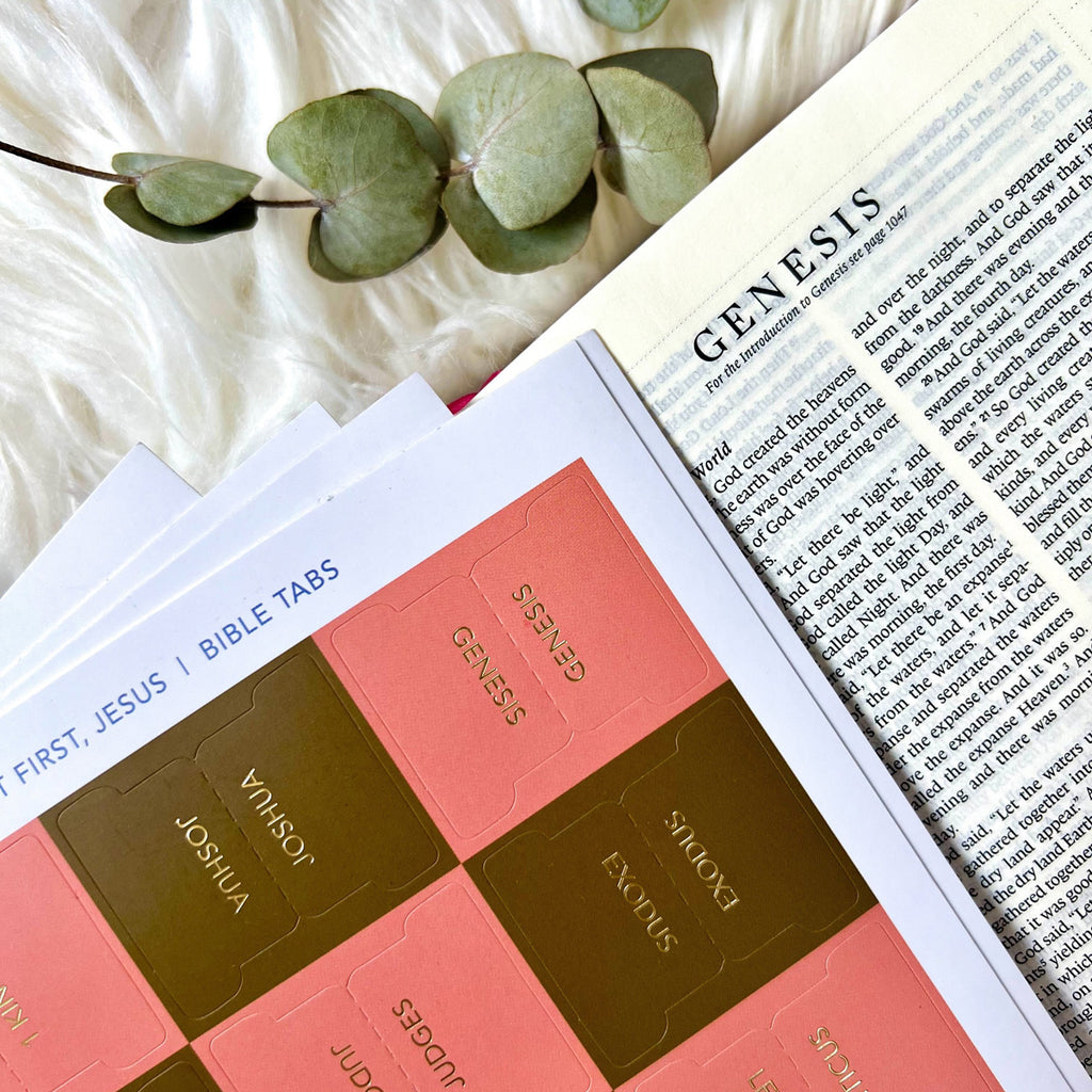 Pink and gold Bible tabs labeled Genesis, Exodus, and Joshua placed on an open Bible page of Genesis, with eucalyptus leaves on a soft white surface.