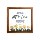A wooden-framed wall art featuring the Bible verse "Put on Love" from Colossians 3:14. The design includes a vibrant arrangement of yellow flowers and green leaves, symbolizing perfect unity and love. Ideal for Christian home decor and inspiration.