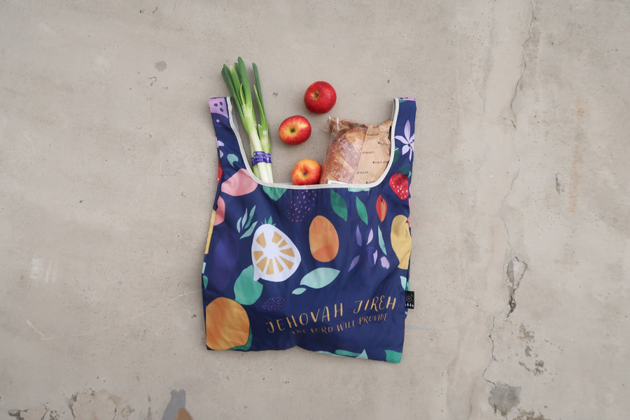 Foldable reusable 
shopping bag with Bible 
text "Jehovah Jireh" and 
colorful fruit design, filled 
with groceries, perfect for 
Christian gifts from a 
Christian gift store.