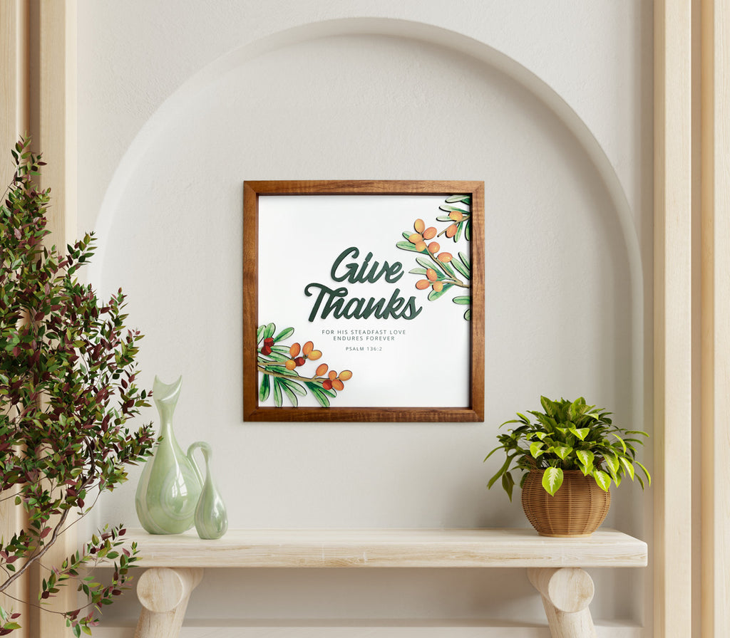 A square wooden-framed wall art featuring the Bible verse "Give Thanks for His Steadfast Love Endures Forever" from Psalm 136:2. The artwork includes vibrant orange berries and green foliage, creating a warm and inviting design. It is displayed in an arched niche with a rustic wooden table adorned with a woven basket containing a lush green plant and elegant glass vases. The setting exudes a peaceful and uplifting atmosphere, perfect for home decor or a meaningful gift.