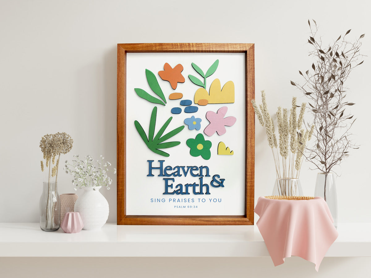 A beautifully crafted wooden poster featuring the Bible verse "Heaven & Earth, Sing Praises to You - Psalm 69:34." The design includes colorful floral and abstract cutouts, displayed on a white shelf adorned with minimalist vases and dried flowers for a modern and serene aesthetic.