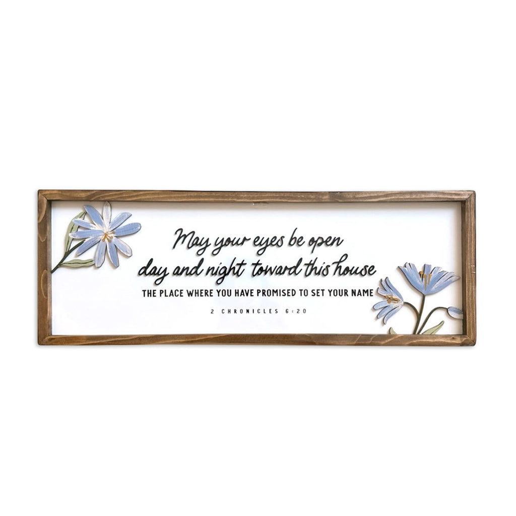 A framed Christian wall art featuring the Bible verse 'May your eyes be open day and night toward this house' from 2 Chronicles 6:20. The design includes delicate blue flowers on both sides, with a rustic wooden frame surrounding the artwork.