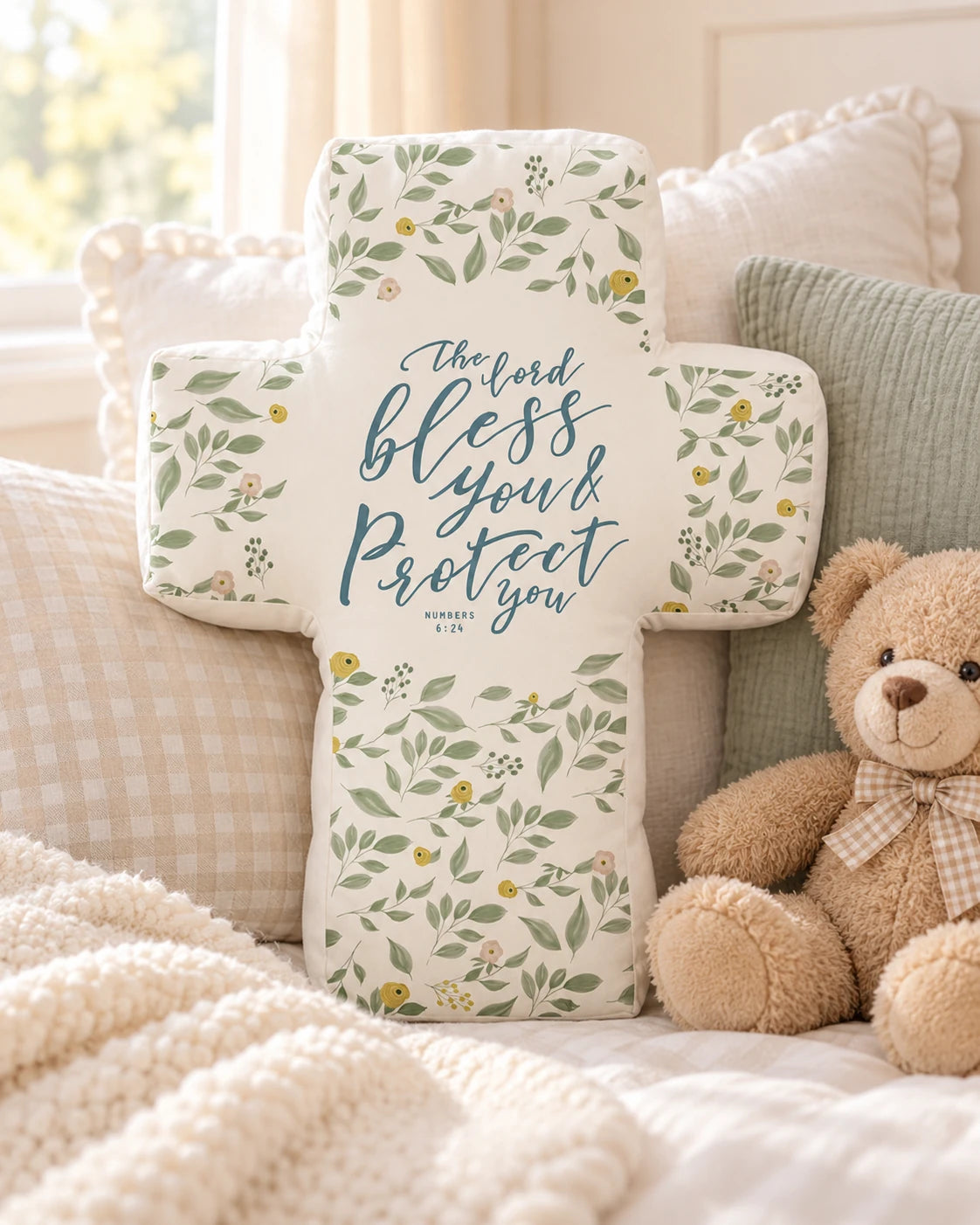 Bless and Protect You floral cross-shaped plush toy styled on a warm cosy bed with pillows