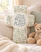Bless and Protect You floral cross-shaped plush toy styled on a warm cosy bed with pillows