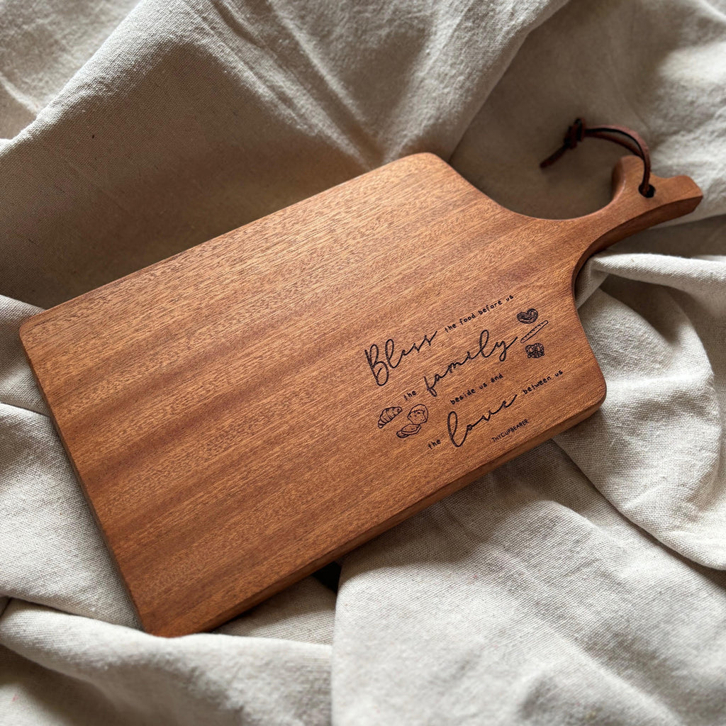 A rustic wooden cheese board with an elegant engraved message: "Bless the food before us, the family beside us, and the love between us." The board features a leather strap and small engravings of bread and hearts, styled on a soft beige fabric background for a warm and cozy aesthetic. Perfect as a kitchen decor piece or for serving.