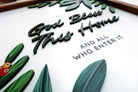 Close-up of a handcrafted wooden scripture poster with the phrase "May God Bless This Home and All Who Enter It." The design features layered text and decorative green leaves, creating a faith-inspired home decor piece.
