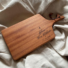 A beautifully crafted wooden cheese board engraved with the phrase "Bless this kitchen and all who enter." The design includes a small wheat symbol and a leather strap for hanging. Styled on soft beige fabric, this rustic board is perfect for serving or as a thoughtful kitchen decor piece.