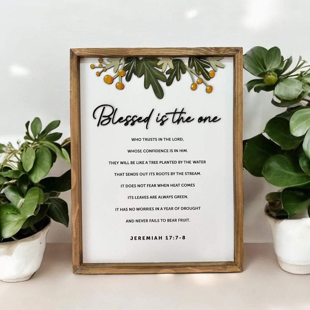 Framed Bible verse wall art featuring Jeremiah 17:7-8 with the phrase 'Blessed is the one,' decorative greenery, and orange accents, surrounded by potted plants.