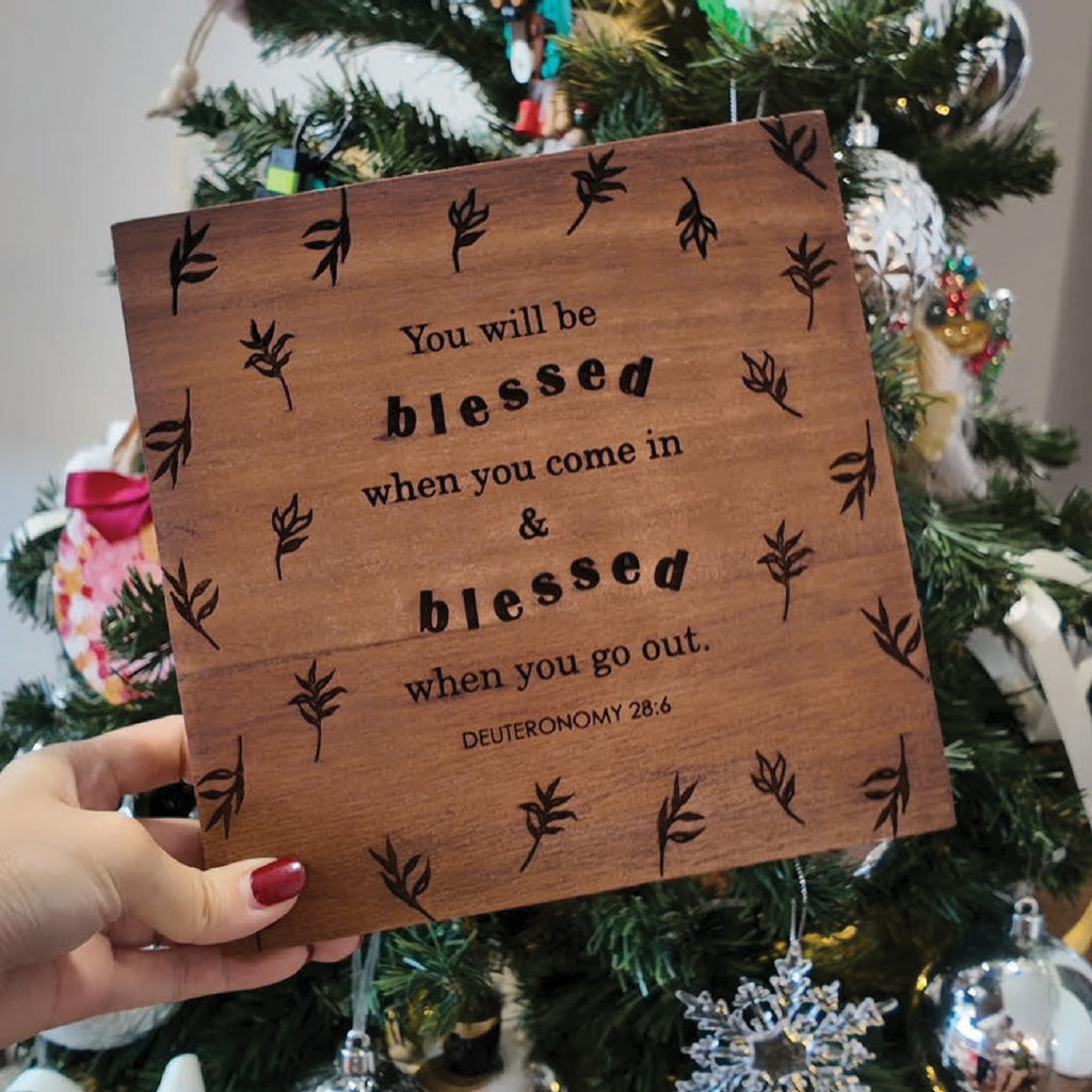 Wooden board decor engraved with the scripture 'You will be blessed when you come in and blessed when you go out. Deuteronomy 28:6,' displayed against a Christmas tree background.