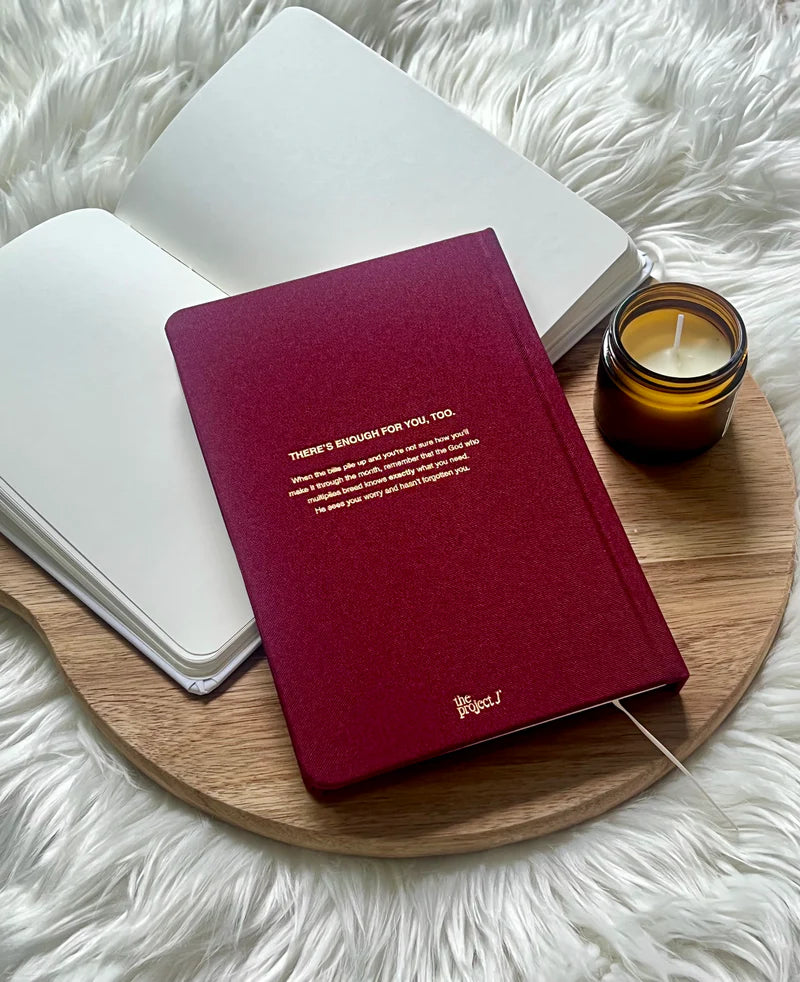 Red fabric journal with gold quote on back cover by Project J placed on wooden tray beside open notebook and candle.