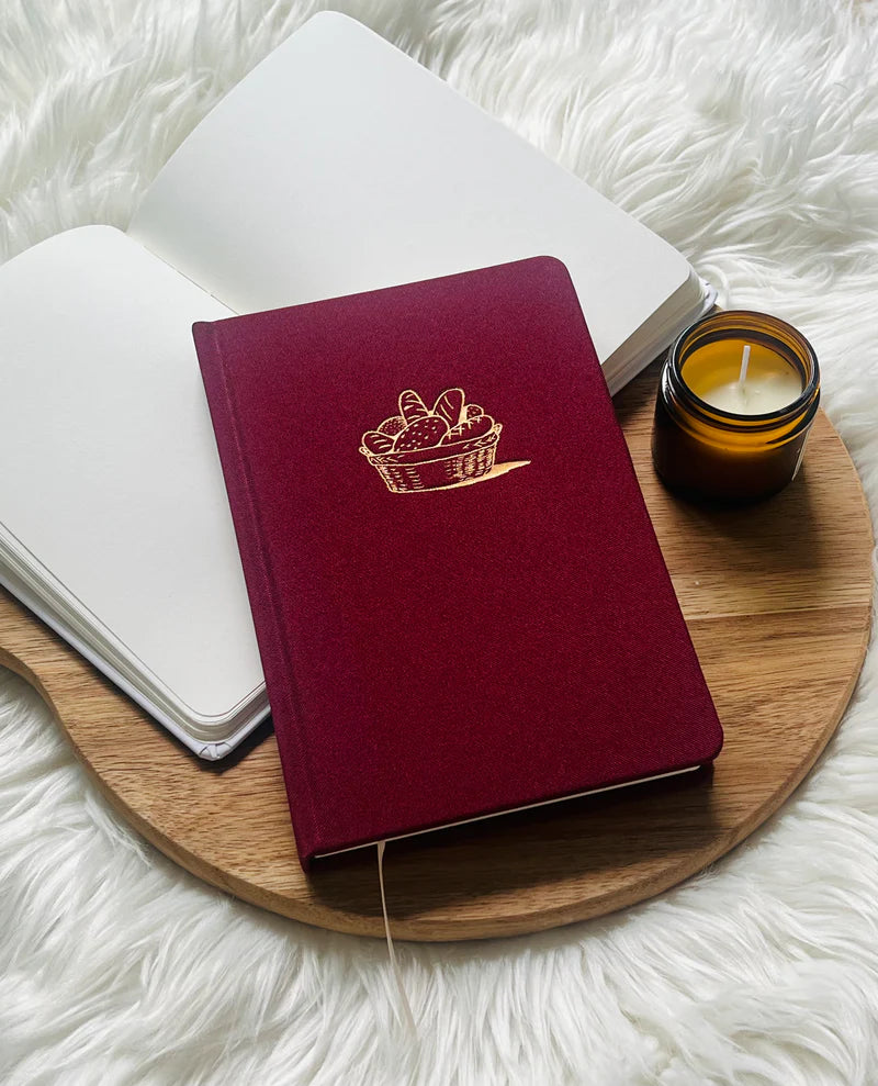 Red fabric journal with gold bread basket design by Project J placed on wooden tray beside open notebook and candle.