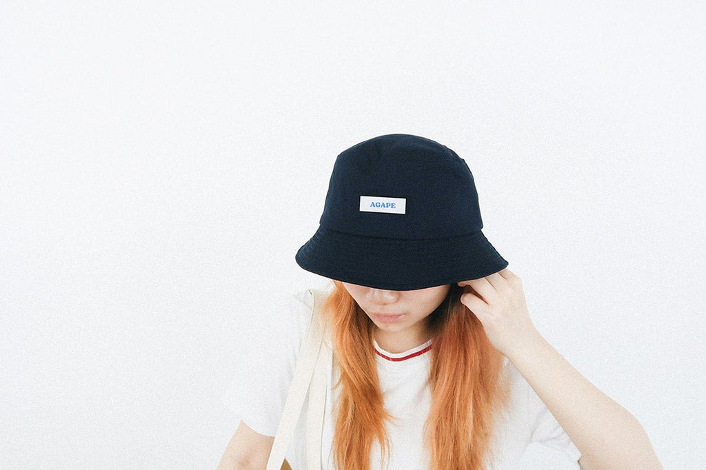 A person wearing a navy blue bucket hat featuring a white patch with the word "AGAPE" in blue, showcasing a faith-inspired and customizable streetwear accessory.