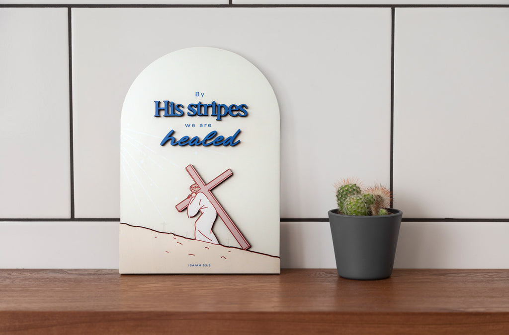 3D wooden Christian wall decor with the phrase 'By His stripes we are healed,' featuring an illustration of Jesus carrying the cross and the Bible verse Isaiah 53:5, displayed on a wooden shelf beside a potted cactus.