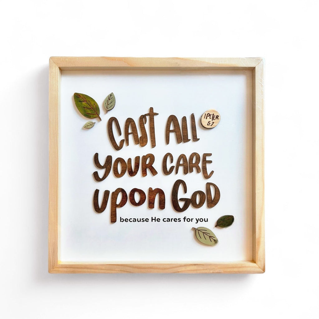 A framed Christian wall art featuring the Bible verse 'Cast all your care upon God because He cares for you' from 1 Peter 5:7. The design includes bold brown text with decorative leaves surrounding the quote, all encased in a minimalist wooden frame.