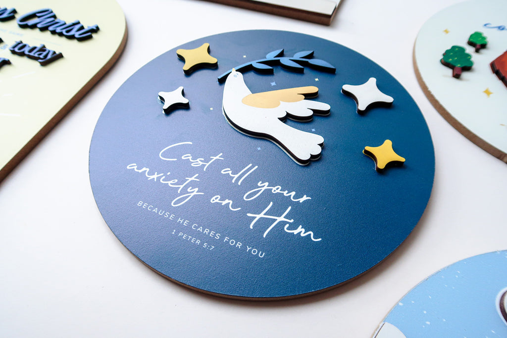 Close-up of a 3D wooden Christian poster with a dove holding an olive branch, surrounded by stars, featuring the Bible verse 'Cast all your anxiety on Him' from 1 Peter 5:7.