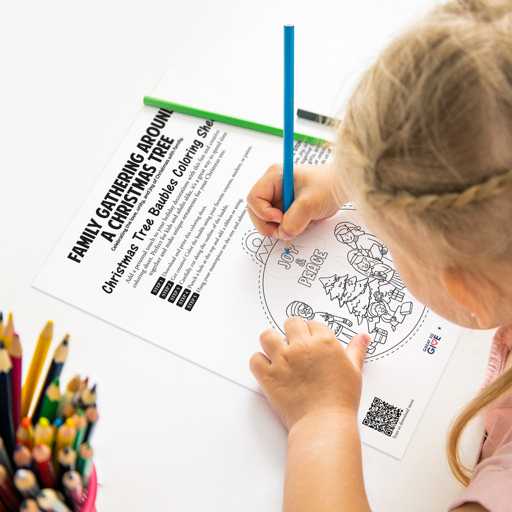 A young child coloring a Christmas bauble activity sheet titled 'Joy & Peace,' featuring a family gathered around a Christmas tree with gift boxes.