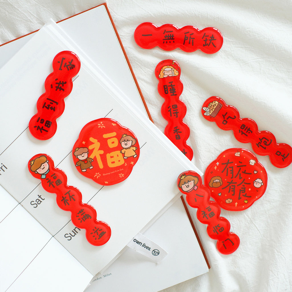 Chinese New Year couplet magnets in red with cute designs featuring blessings and prosperity messages, arranged on a white planner.