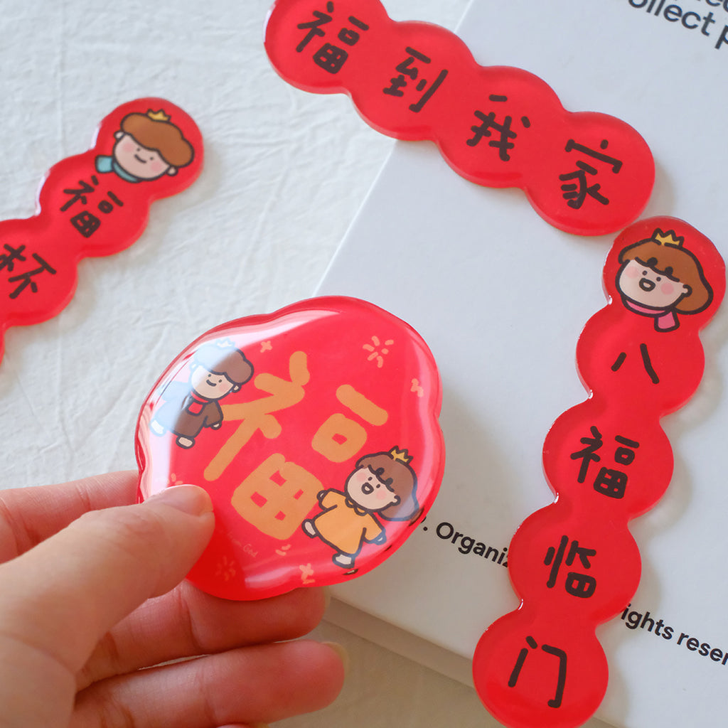 Chinese New Year couplet magnets featuring red blessings and cute character designs, held by a hand on a white background