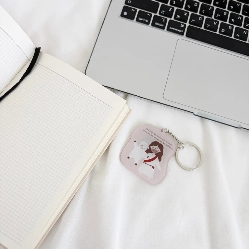 God Loves Us Christian acrylic keychain styled beside a notebook and laptop for everyday use