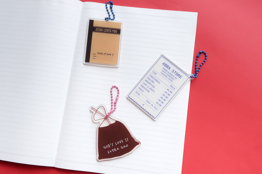 Christian acrylic keychains styled on an open notebook featuring Jesus Loves You jotter book, Abba Store receipt, and God’s Love Is Extra Gao drink bag designs