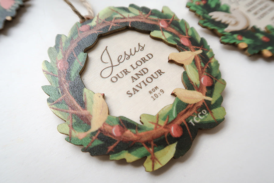 Close-up of a Christian Christmas wreath ornament featuring the Bible verse Romans 10:9 with birds and holly decorations.