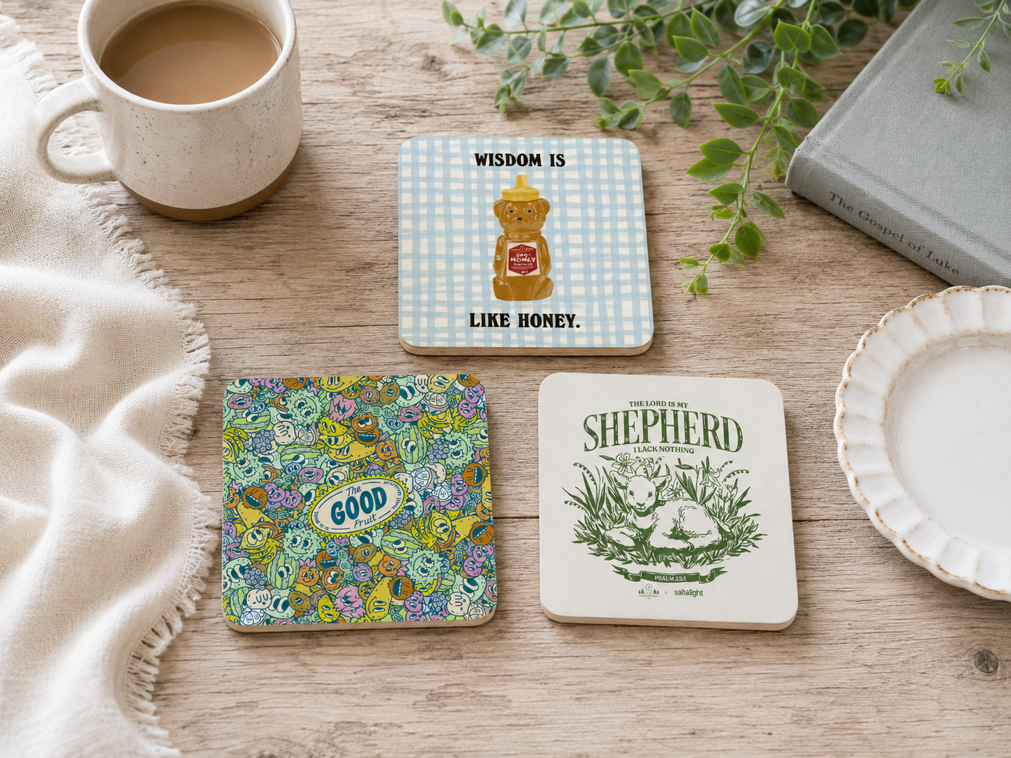 Christian coaster set featuring The Good Fruit, Wisdom Is Like Honey, and The Lord Is My Shepherd designs styled on a wooden table with a mug, Bible, greenery, and plate.