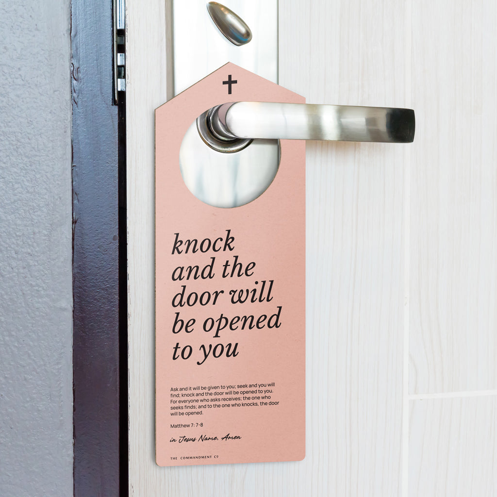 Peach-colored Christian door hanger with the message 'Knock and the door will be opened to you' from Matthew 7:7-8, hanging on a modern silver door handle attached to a light wood door.