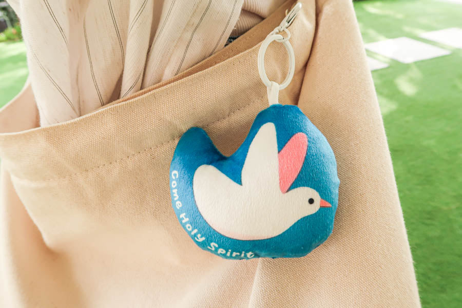 Blue plush keychain with a white dove design and 'Come Holy Spirit' message, clipped onto a beige bag as a symbol of peace and faith.