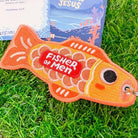 Orange embroidered fish-shaped keychain with the message 'Fisher of Men' in red, displayed on vibrant green grass with Christian-themed cards in the background.