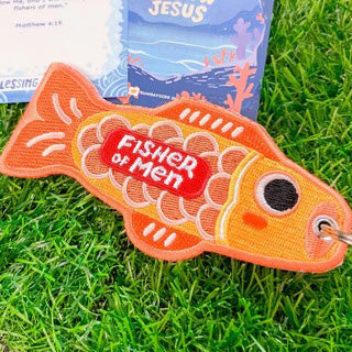 Orange embroidered fish-shaped keychain with the message 'Fisher of Men' in red, displayed on vibrant green grass with Christian-themed cards in the background.