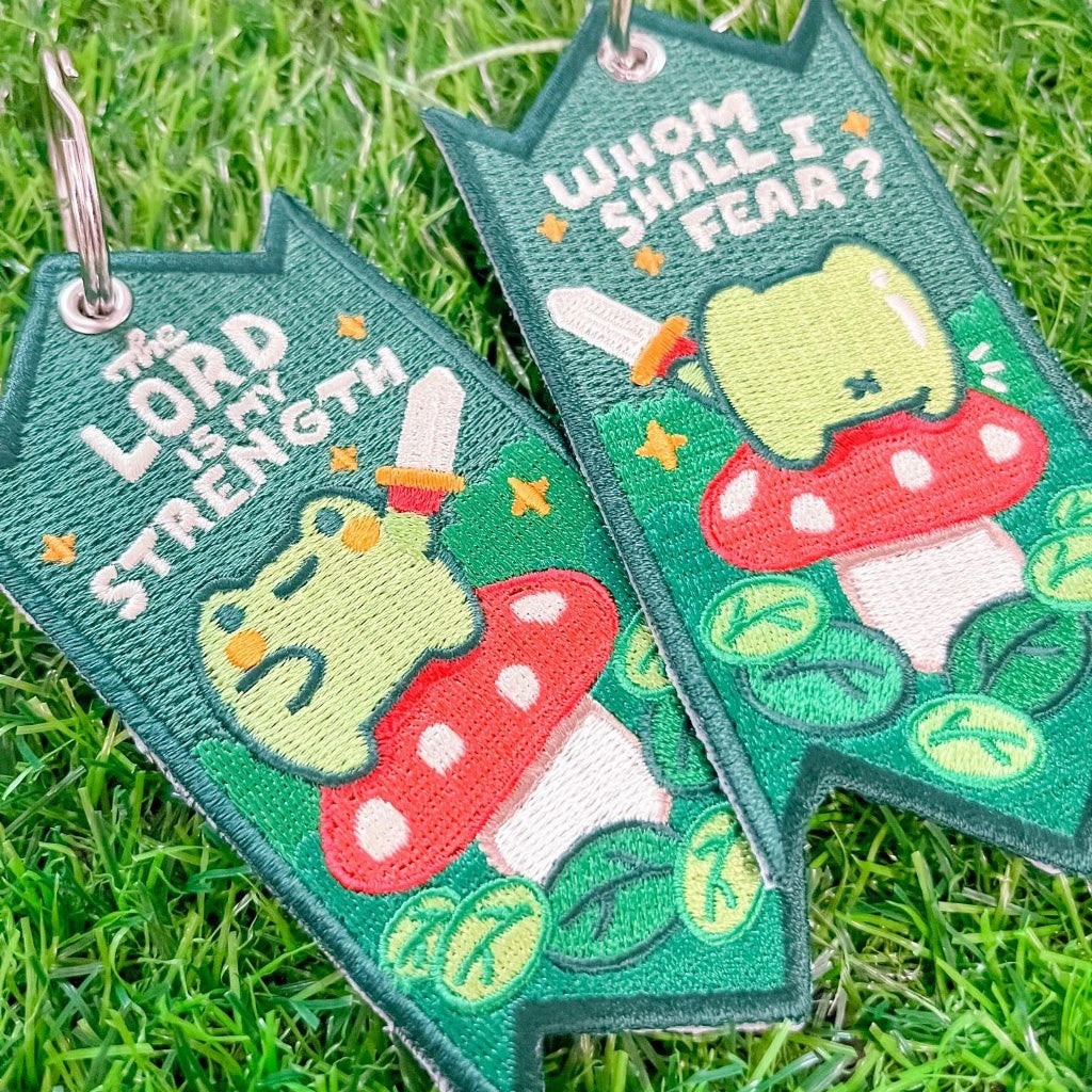 Close-up of two embroidered Christian keychains featuring a frog sitting on a red mushroom, with swords and the messages 'The Lord is My Strength' and 'Whom Shall I Fear?' displayed on a grassy background.